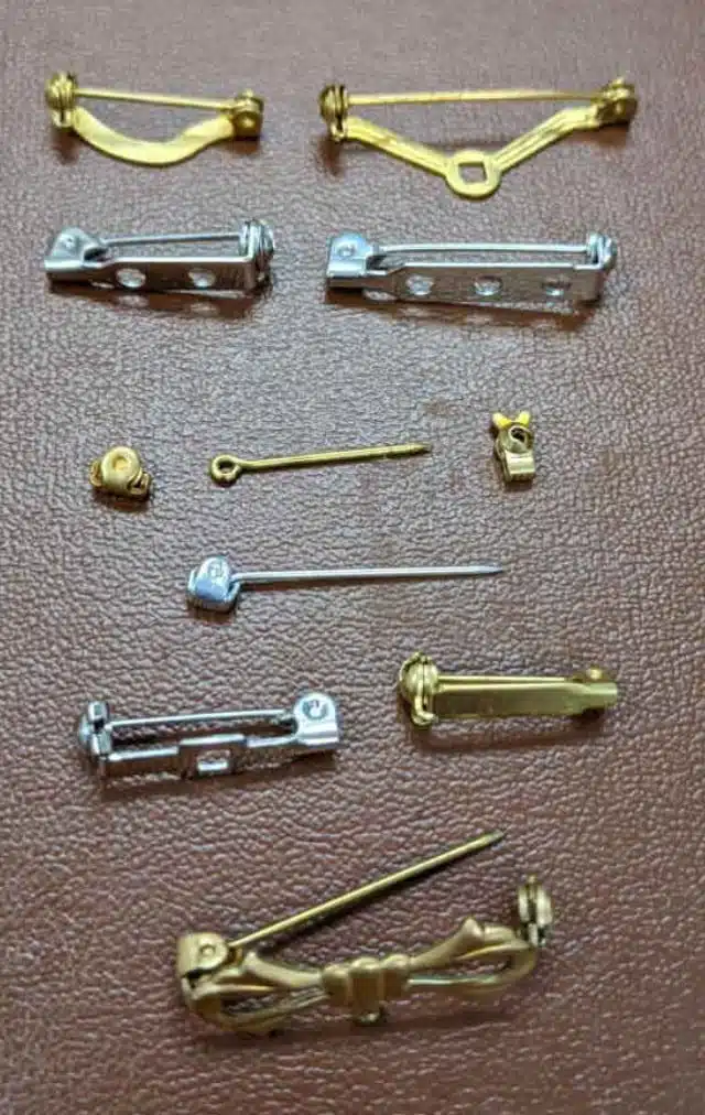 Assortment of small gold and silver metal brooch pins and clasps laid on a brown surface, showing various fasteners like T-bar, C-clasps, and pin stems.