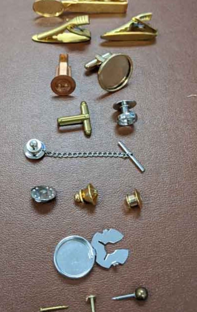 Collection of assorted small metal hardware pieces (cufflinks, buttons, tie tacks) laid out on a brown surface, showing various shapes and finishes in gold and silver tones.