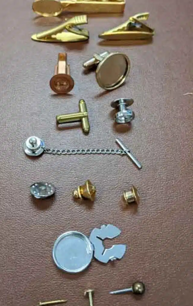 Collection of assorted small metal hardware pieces (cufflinks, buttons, tie tacks) laid out on a brown surface, showing various shapes and finishes in gold and silver tones.