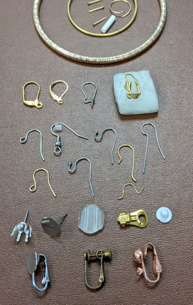 Assorted jewelry findings and components laid out on a brown surface: earring hooks, clasps, jump rings, and decorative pieces in gold, silver, and copper tones.