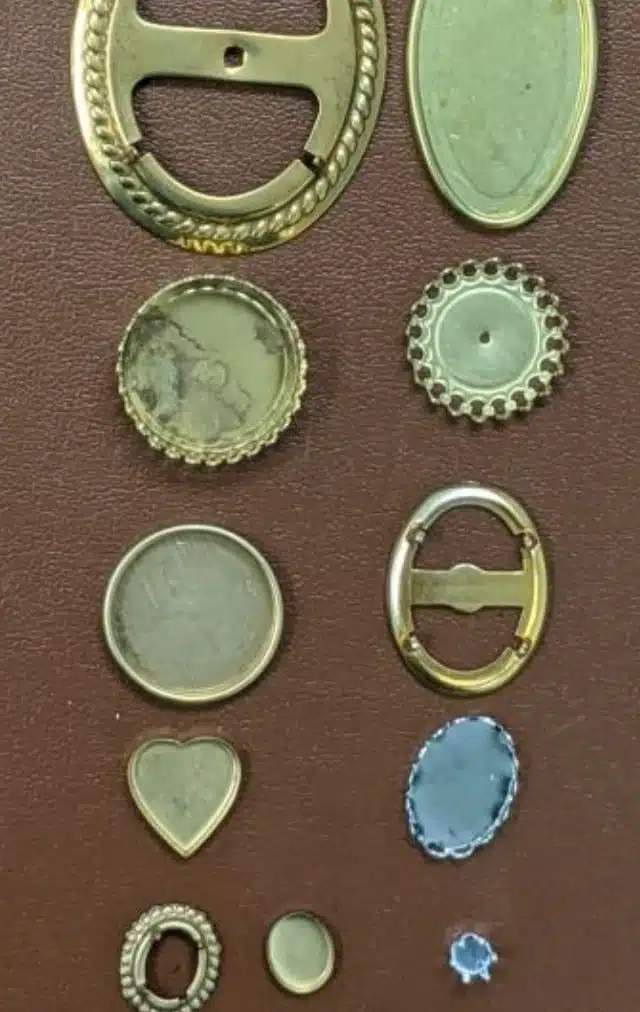 A collection of small metal findings and buttons laid out on brown leather, including buckles, gear-shaped caps, a heart-shaped charm, and a blue cabochon setting.