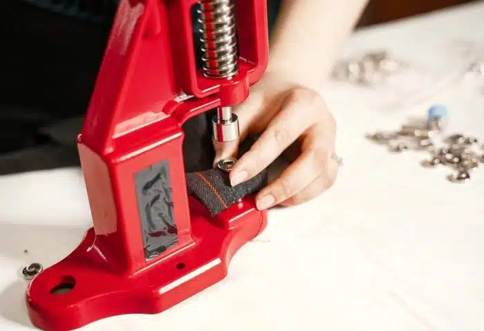 Person using a red snap fastener tool to attach a metal snap to dark fabric on a work table, with loose snaps nearby